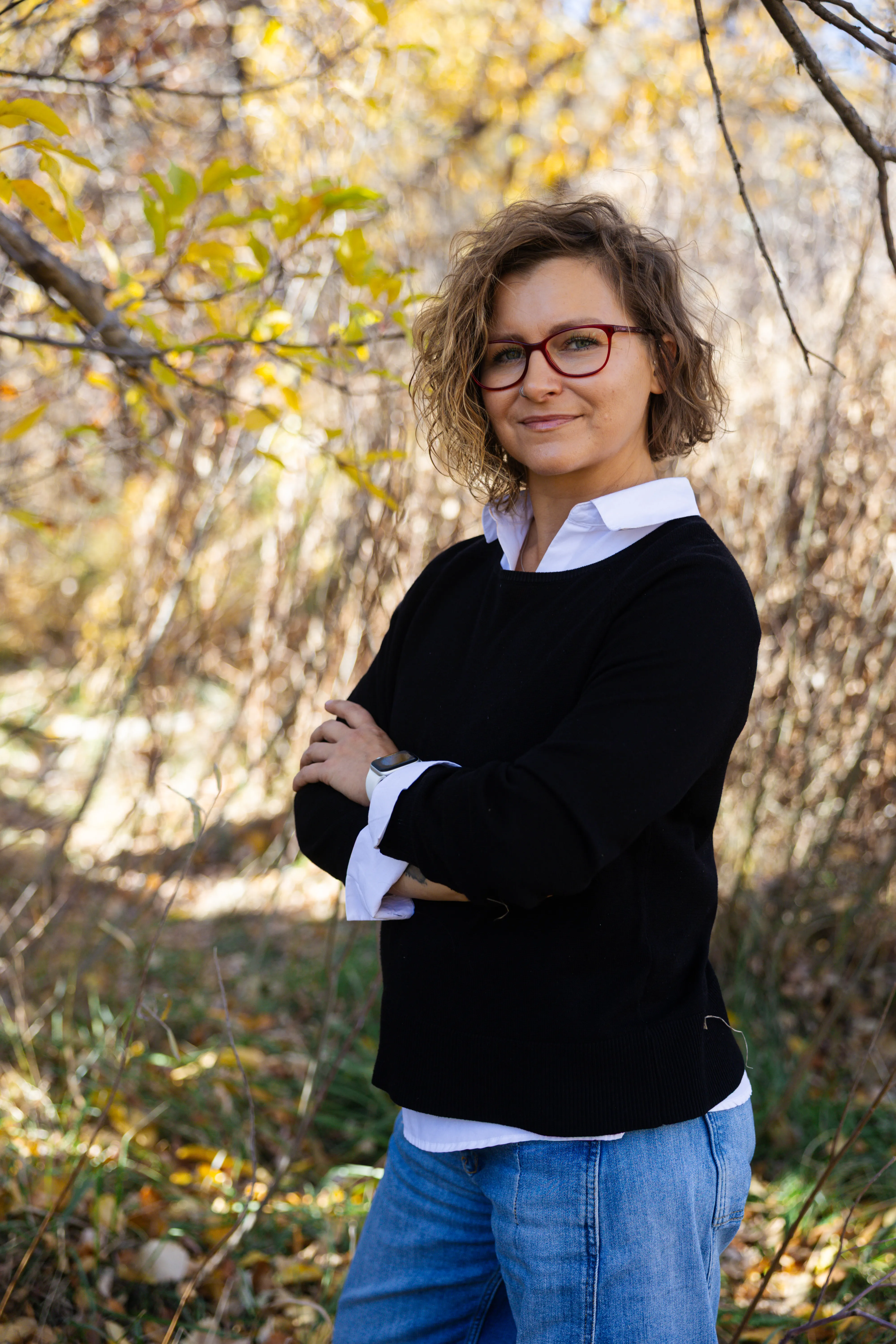 Varvara Lytkina standing among autumn cottonwoods in Colorado, arms crossed, wearing a black sweater and red glasses